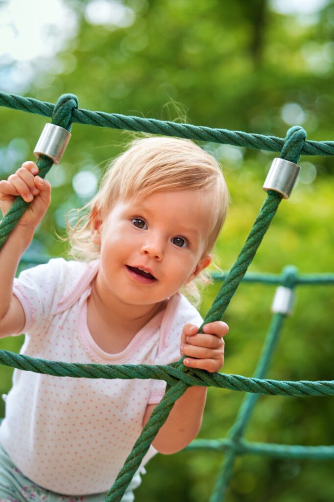 Fröhliches Kind blickt zwischen Seilen hindurch und lacht, sommerliches Grün im Hintergrund.