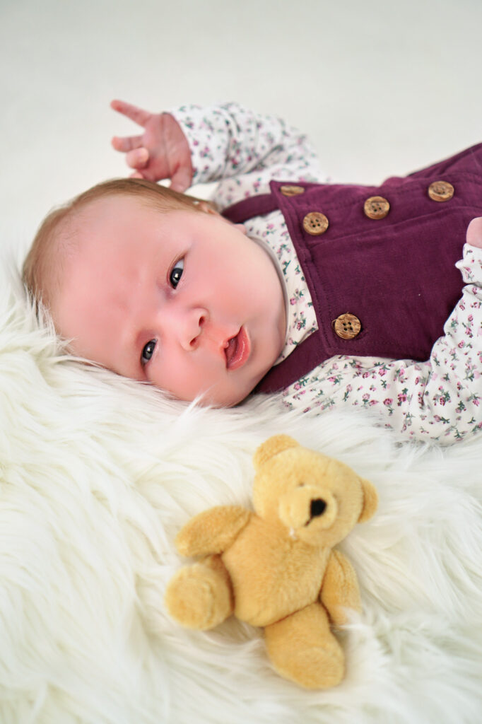 Baby liegt auf weißem Fell neben einem kleinen Teddybär – Newbornfoto von Herzberger Fotografie