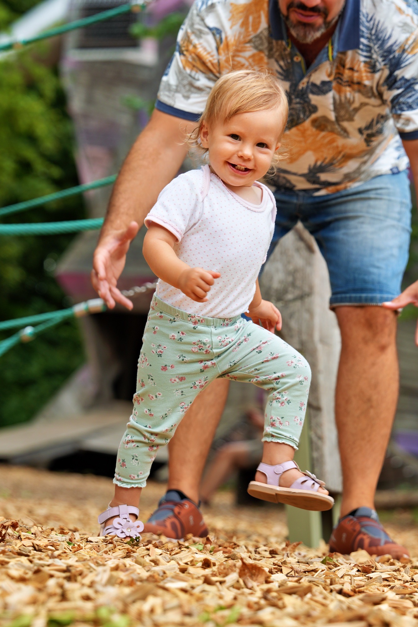Kleinkind läuft lachend über Holzhackschnitzel, der Vater sichert im Hintergrund. - Familienfotografie