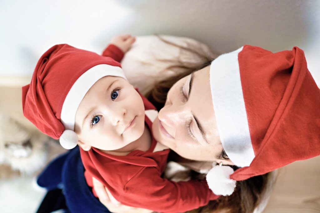 Mutter h&auml;lt ihr Baby im Arm, beide tragen rote Nikolausm&uuml;tzen und l&auml;cheln in die Kamera