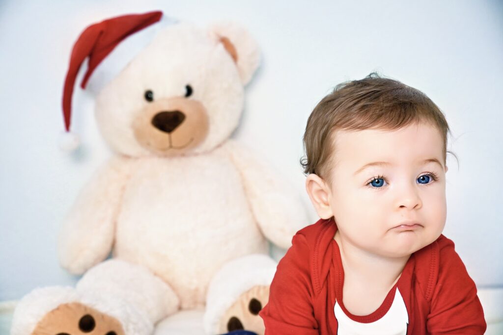 Baby mit roter Nikolausmütze sitzt vor einem großen Teddybären und schaut neugierig in die Kamera