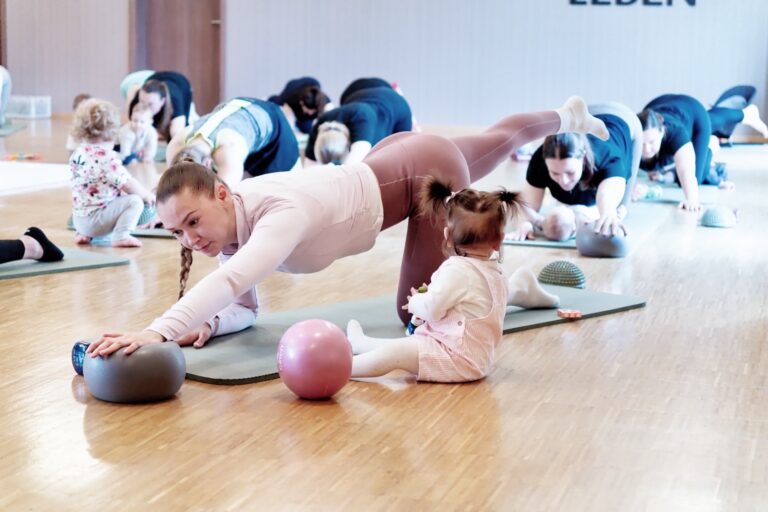 Mutter im Unterarmst&uuml;tz mit erhobenem Bein, ihr Baby sitzt direkt davor auf der Matte und schaut zu ihr hoch