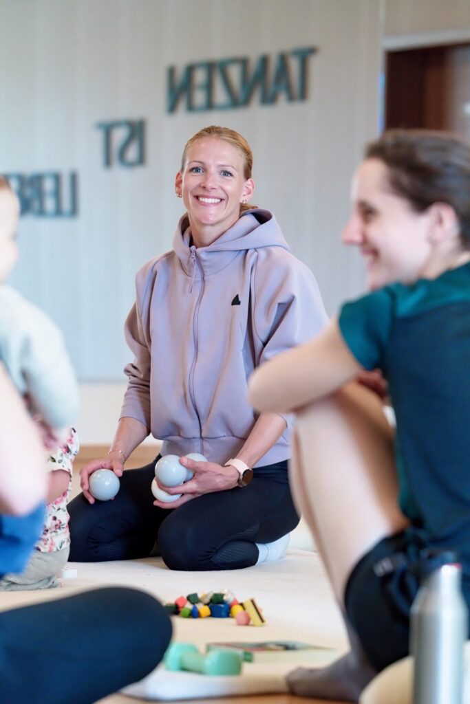 Kimberly Müller sitzt auf einer Sportmatte und hält zwei Faszienrollen, umgeben von Kursteilnehmerinnen im Hintergrund