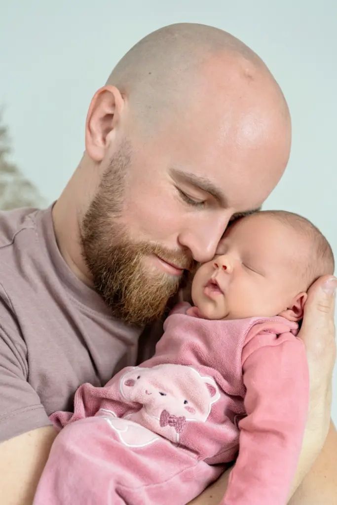 Vater mit Bart h&auml;lt sein schlafendes Baby im rosa Strampler an seine Wange gekuschelt, die Augen geschlossen