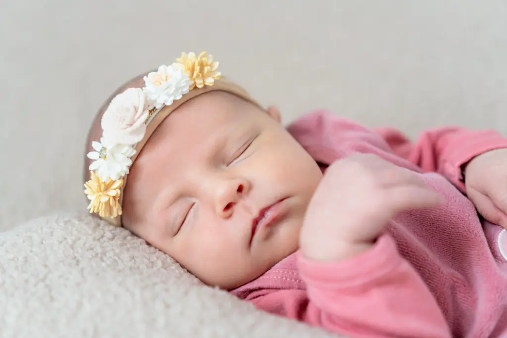 Schlafendes Baby mit cremefarbenem Blumen-Haarband liegt auf einem beigen Kissen, die Hand am Kinn