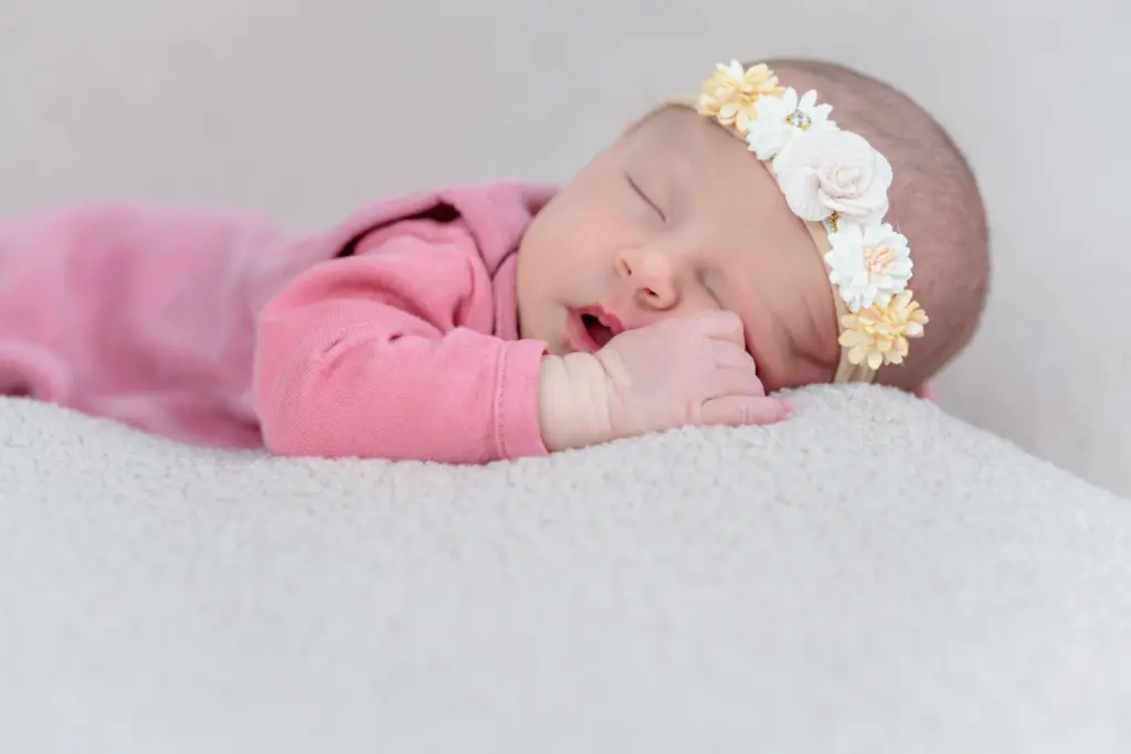 Baby mit Blumen-Haarband in rosa Strampler schl&auml;ft tief, Kopf und Arm ruhen auf einem hellen Kissen