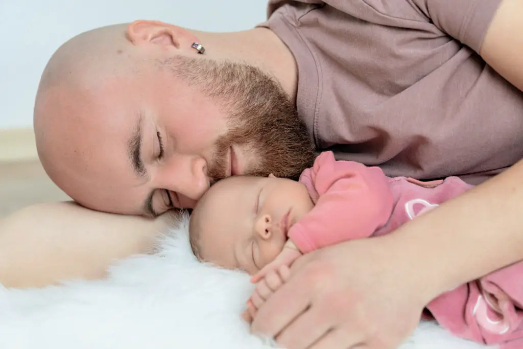 Vater liegt mit geschlossenen Augen neben seinem schlafenden Baby auf einem wei&szlig;en Fell, die Wange an das Baby gelegt
