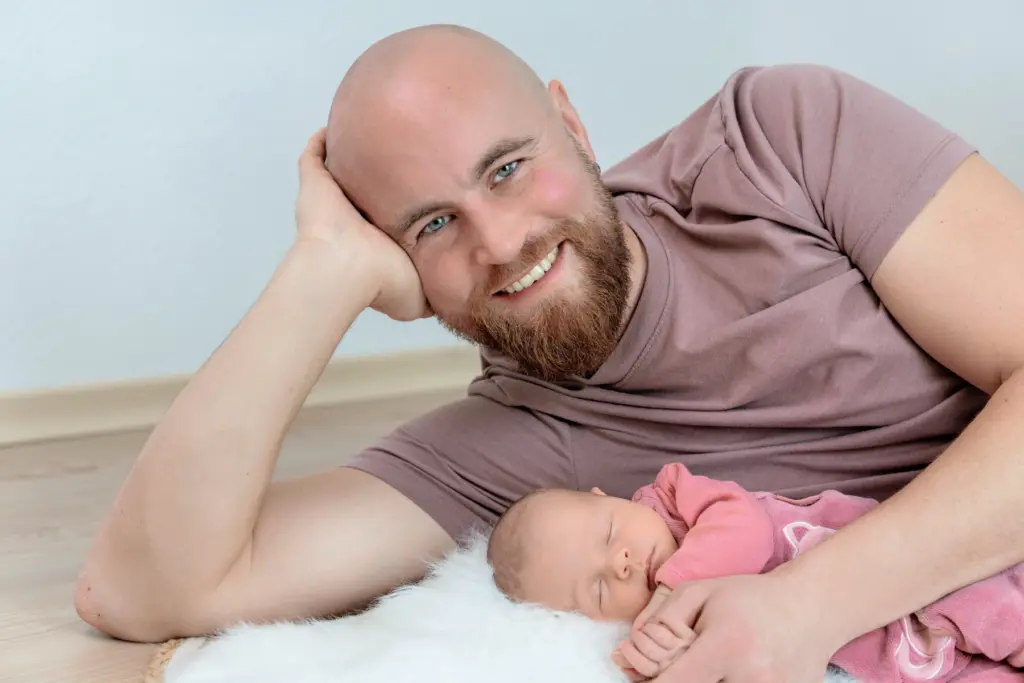 L&auml;chelnder Vater mit Bart, Kopf in die Hand gest&uuml;tzt, liegt hinter seinem Baby im rosa Strampler auf wei&szlig;em Fell