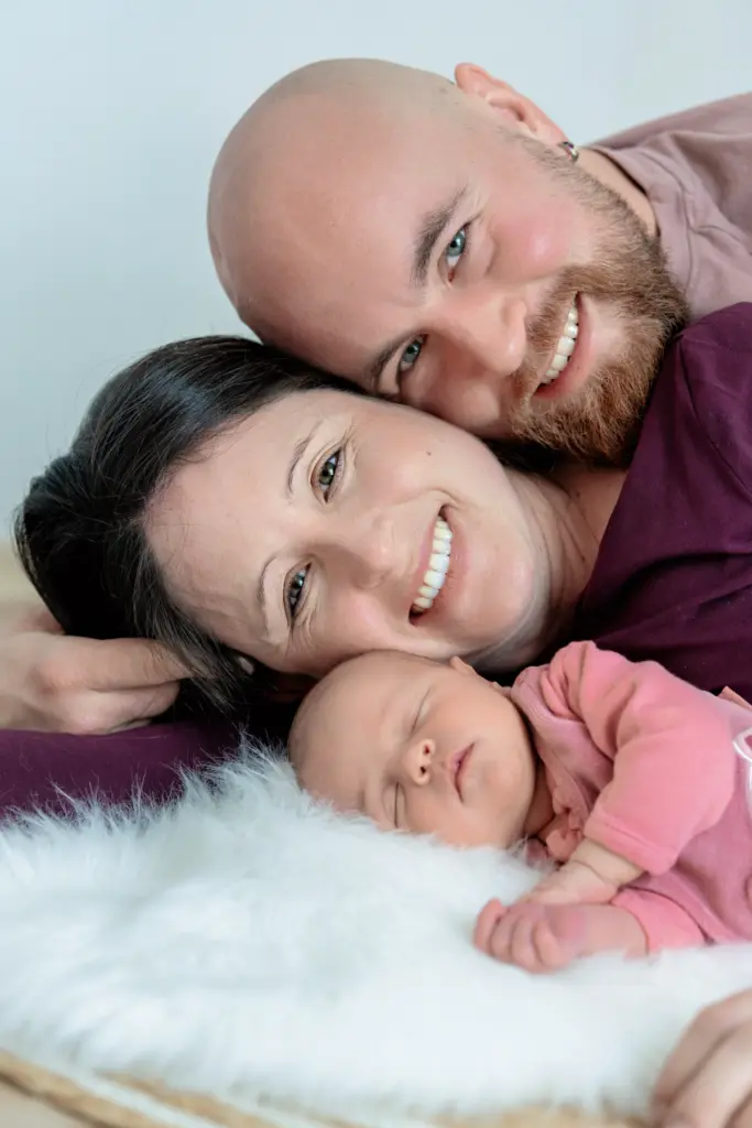 Lachende Eltern schmiegen die Wangen aneinander, unter ihnen liegt ihr Baby auf einem wei&szlig;en Fell