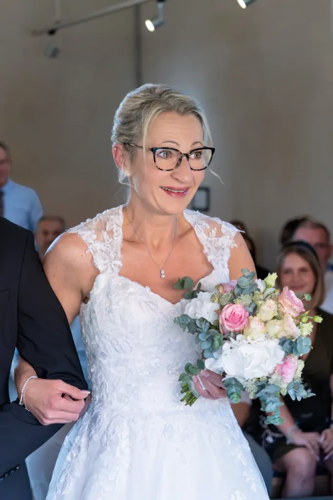Braut in Spitzenkleid geht mit Brautstrau&szlig; am Arm eines Mannes im dunklen Anzug zur Trauung, G&auml;ste im Hintergrund