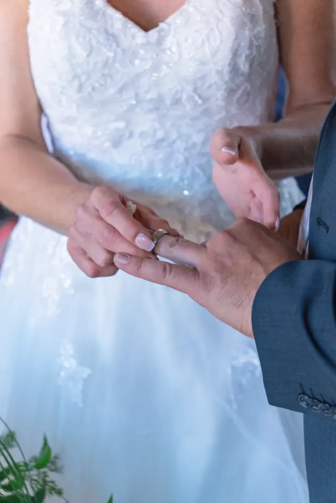 Nahaufnahme: Der Ehering wird der Braut an den Finger gesteckt, im Hintergrund das wei&szlig;e Brautkleid