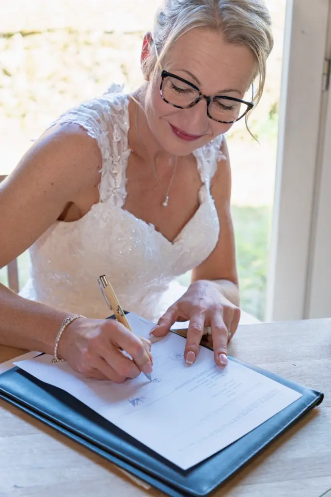 L&auml;chelnde Braut mit Brille unterschreibt an einem Tisch eine Urkunde, den Stift in der Hand