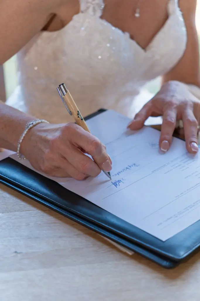 Nahaufnahme einer Hand mit Stift beim Unterschreiben in einer schwarzen Mappe, dahinter das wei&szlig;e Brautkleid