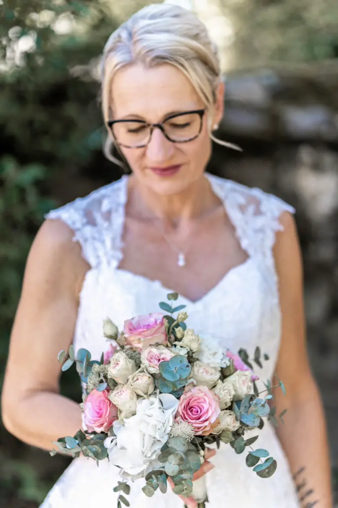 Portr&auml;t einer strahlenden Braut mit Brille im Spitzenkleid, die ihren rosa-wei&szlig;en Brautstrau&szlig; h&auml;lt