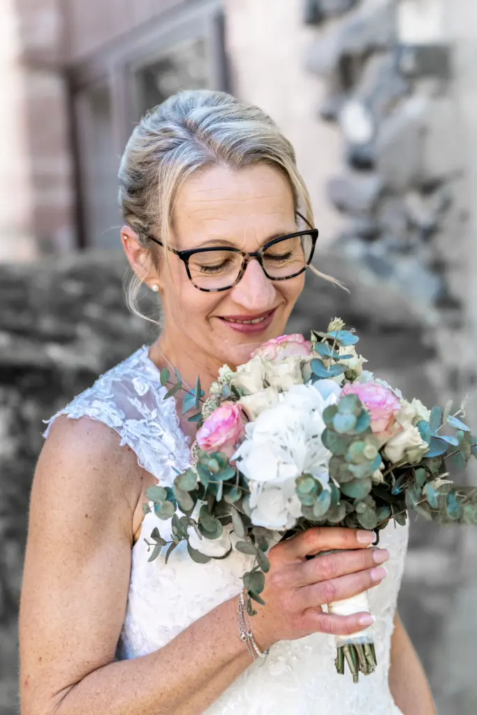 Braut mit Brille schaut l&auml;chelnd auf ihren Brautstrau&szlig; aus rosa und wei&szlig;en Blumen mit Eukalyptus