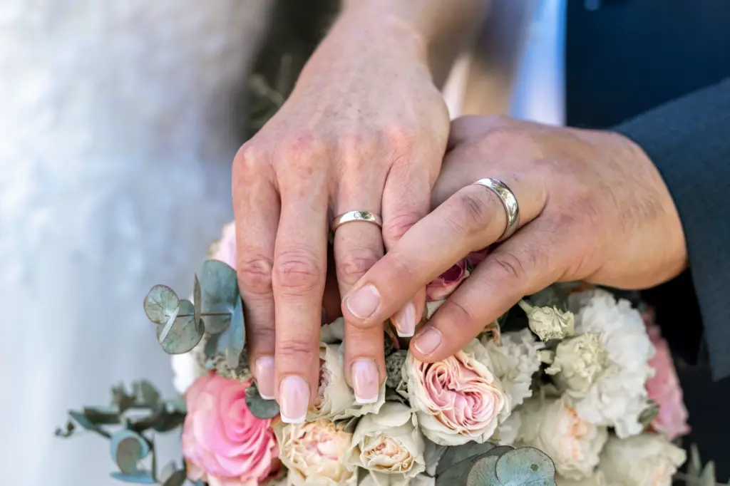 Nahaufnahme zweier H&auml;nde mit Eheringen, die auf einem Brautstrau&szlig; aus rosa und wei&szlig;en Rosen ruhen