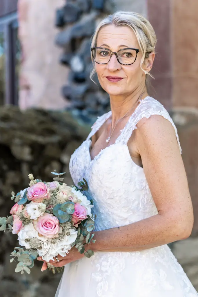 Braut mit Brille im Spitzenkleid steht vor einer Steinmauer und h&auml;lt l&auml;chelnd ihren Brautstrau&szlig;