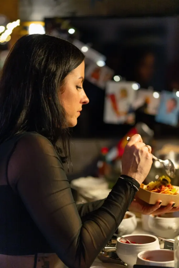 Frau in schwarzem Oberteil mit transparenten &Auml;rmeln nimmt sich Essen vom Buffet, im Hintergrund Lichterkette