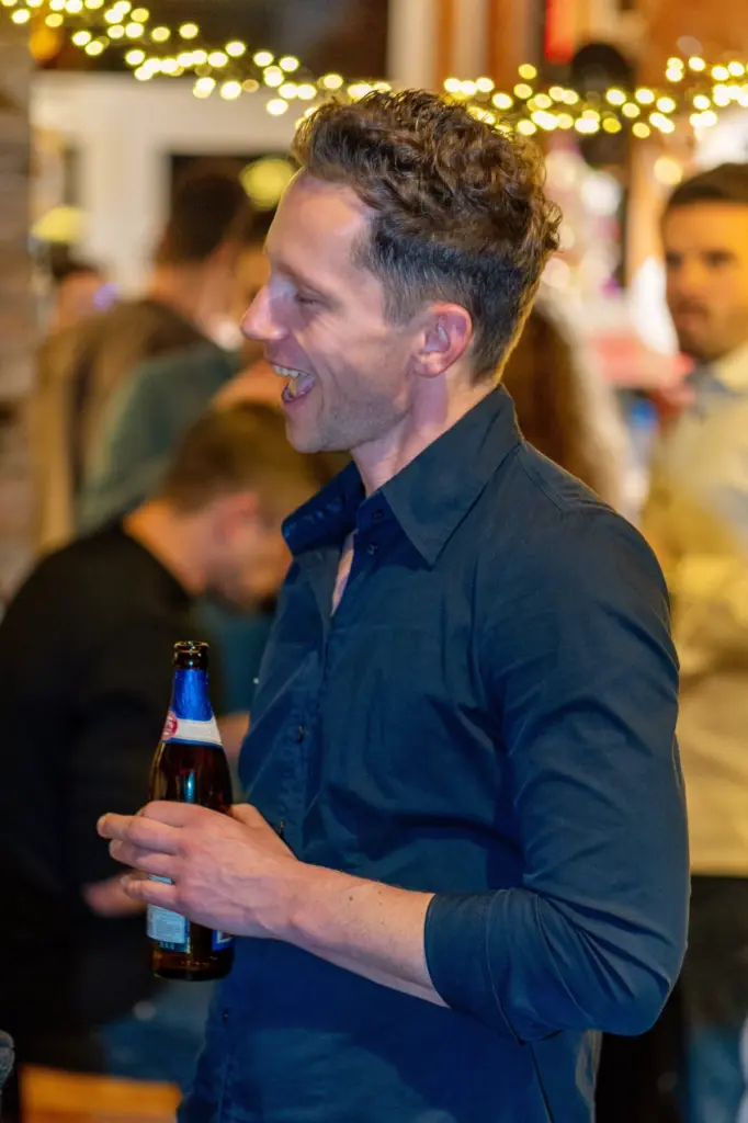 Mann im dunkelblauen Hemd mit Bierflasche in der Hand blickt zur Seite, dahinter G&auml;ste und Lichterkette