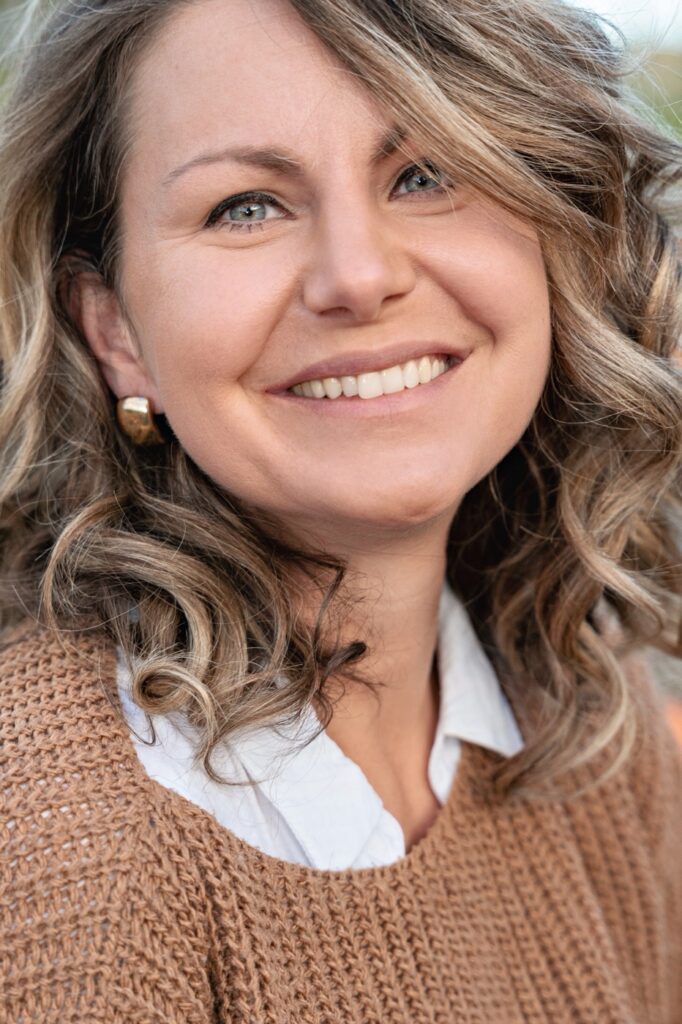 Portr&auml;t von Daria Herzberger: l&auml;chelnde Frau mit welligen, blond-braunen Haaren, braunem Strickpullover und wei&szlig;er Bluse