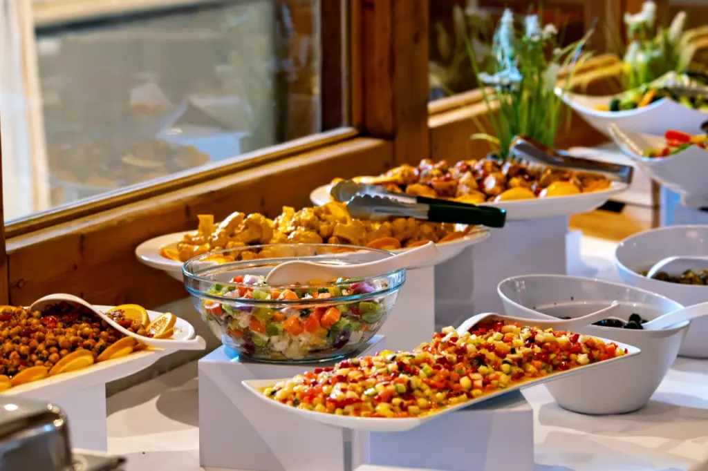 Reich gedecktes Buffet mit Salaten, warmen Gerichten und Servierzange vor einem Holzfenster
