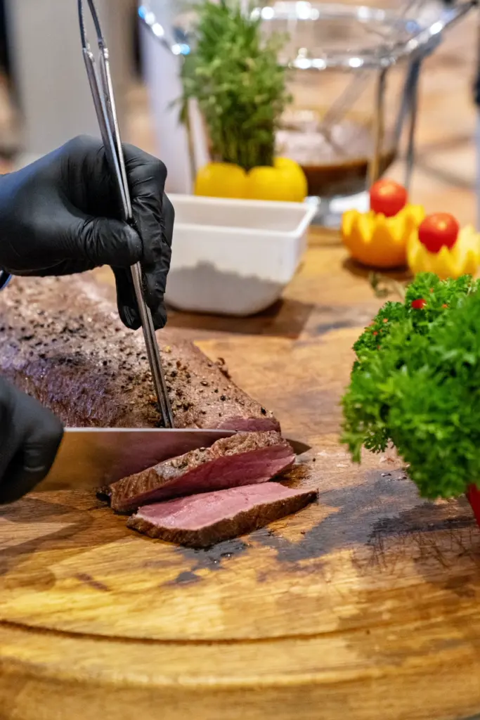 Koch mit schwarzen Handschuhen tranchiert rosa gebratenes Fleisch auf einem Holzbrett am Buffet
