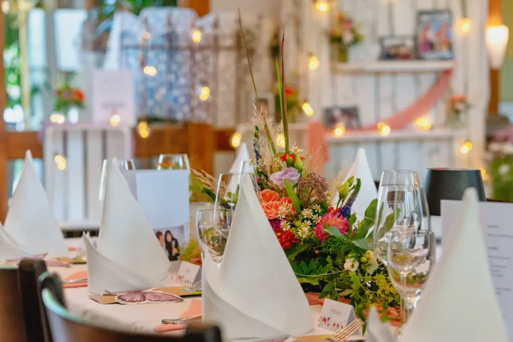 Festlich gedeckter Tisch mit Blumengesteck, wei&szlig;en Servietten und Weingl&auml;sern, im Hintergrund eine Lichterkette