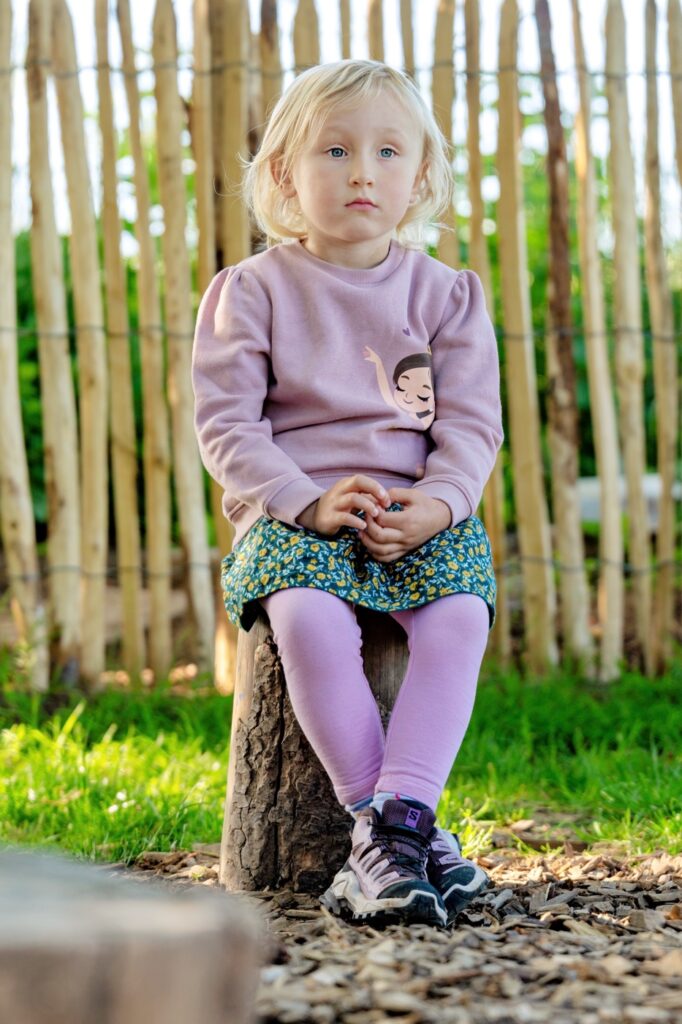 M&auml;dchen in lila Pullover, Rock und Wanderschuhen sitzt auf einem Baumstumpf vor einem Zaun aus Holzst&auml;ben im Kita-Garten