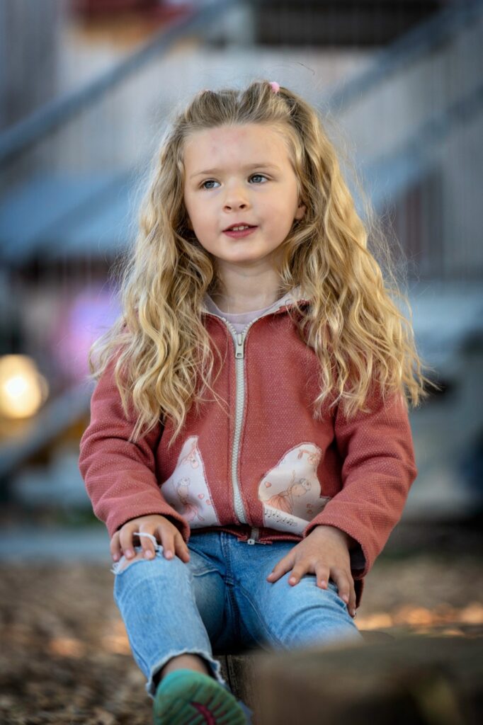 M&auml;dchen in roter Jacke und Jeans sitzt entspannt auf dem Spielplatz und schaut zur Seite, im Hintergrund unscharfe Holzger&auml;te