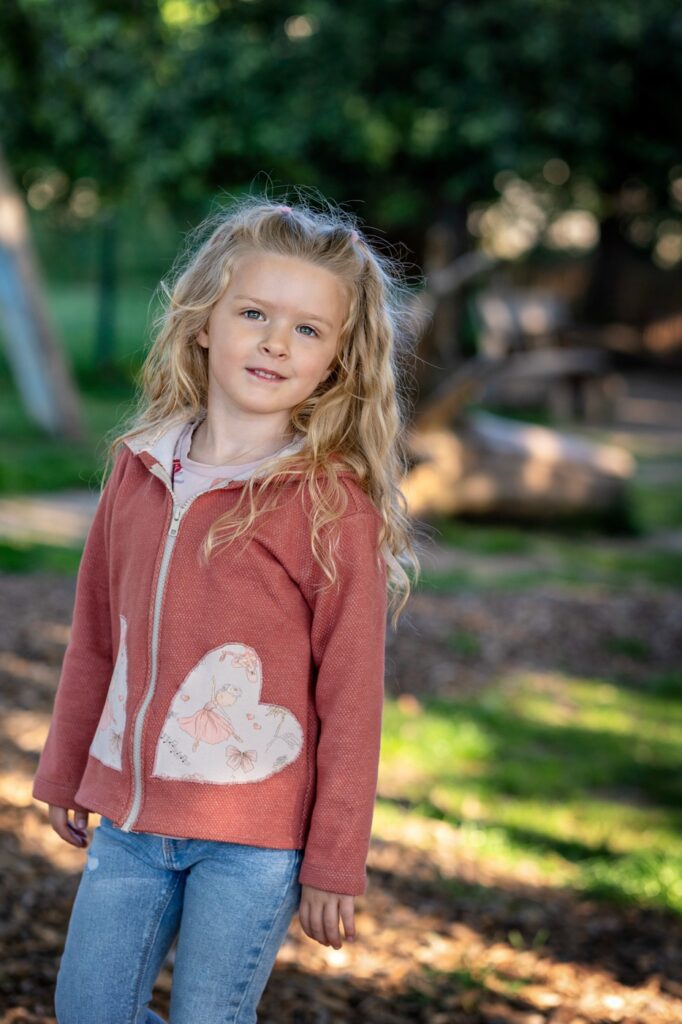 M&auml;dchen in roter Jacke steht auf Holzhackschnitzeln zwischen Baumst&auml;mmen auf einem Naturspielplatz und blickt zur Kamera