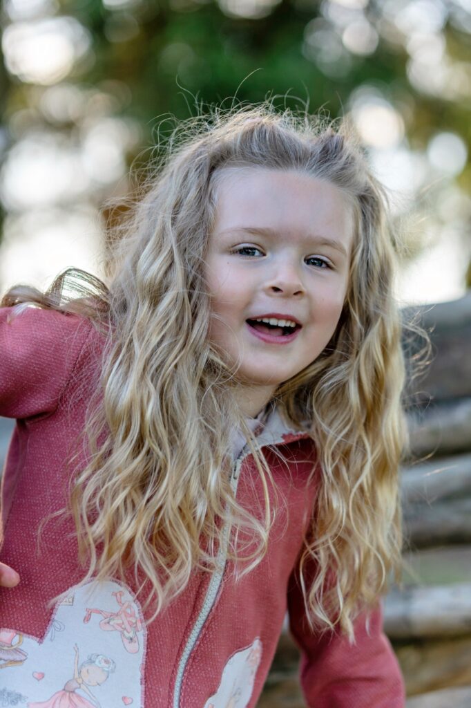 M&auml;dchen mit langen blonden Locken und roter Jacke mit Herz lacht und beugt sich der Kamera entgegen, drau&szlig;en im Gr&uuml;nen